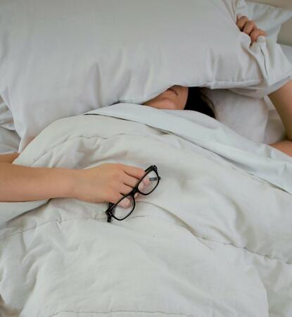 Person lying in bed under white sheets, holding a pair of glasses, with a pillow covering part of their face.