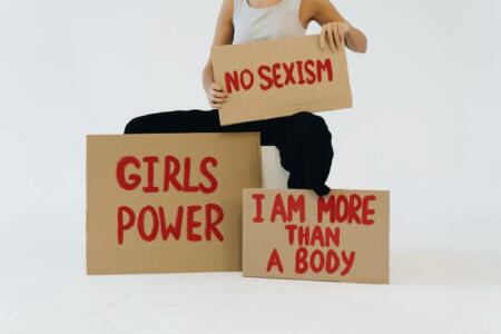 A person holds signs with the messages "GIRLS POWER," "NO SEXISM," and "I AM MORE THAN A BODY.
