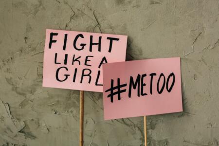 Two pink signs with the messages "FIGHT LIKE A GIRL" and "#METOO" against a wall background.